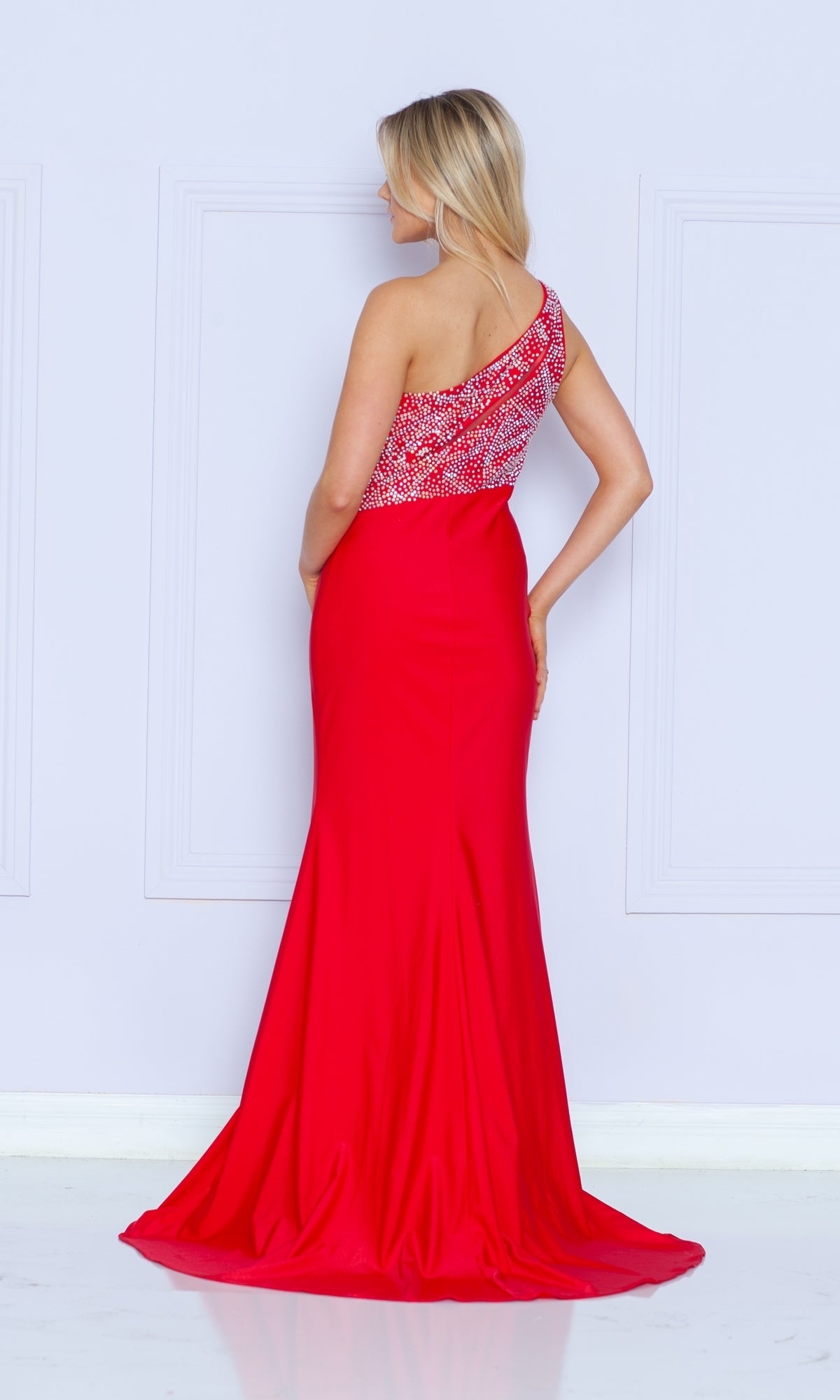 A woman in the Poly USA 9116 One-Shoulder Long Jersey Prom Dress, featuring a red beaded bodice, stands facing away from the camera against a light-colored wall.